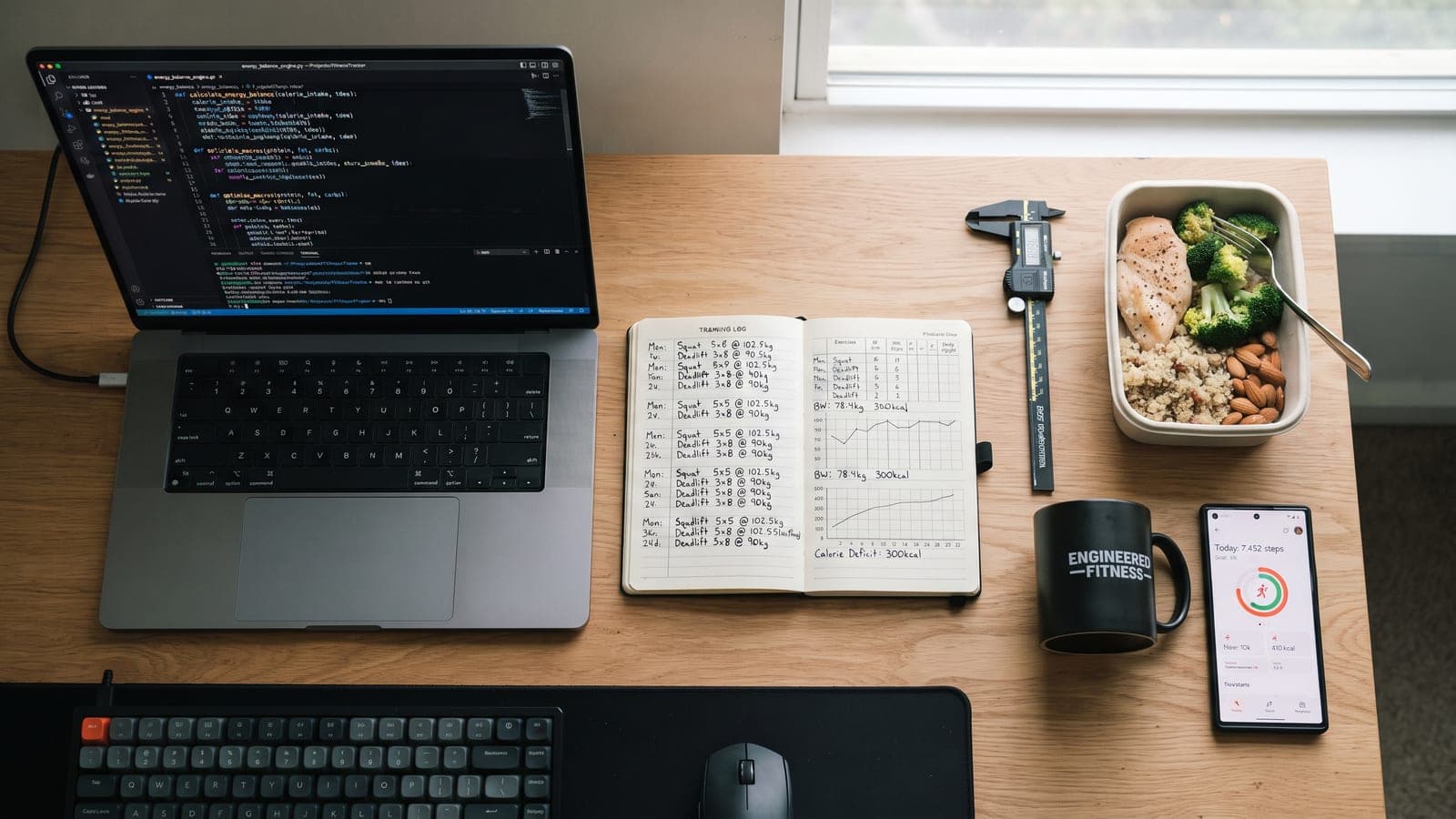 A desk with code on a laptop, a handwritten training log, calipers and a prepared meal